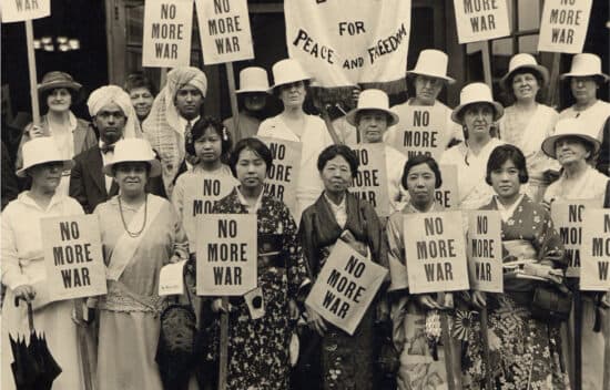 A group of women of different ethnicities and cultural dress stand together and hold signs that read No More War.