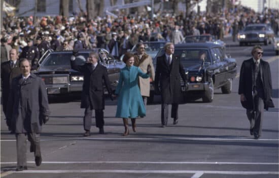 Jimmy Carter and his wife walk on the street in a parade. Cars follow them. A crowd lines the side of the street.