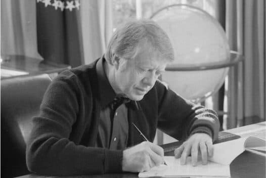 Jimmy Carter sits at a desk and signs a document.