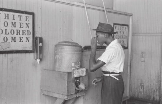 An African American man drinks from a water fountain labeled Colored.