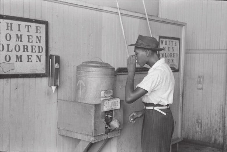 An African American man drinks from a water fountain labeled Colored.