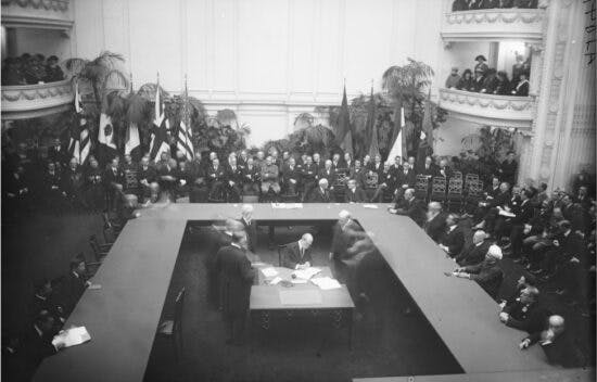 Men sit around a U-shaped table. A group of men sit at a table in the middle and sign some papers. Other men sit around the room and in balconies. Multiple flags are posted at the back of the room.