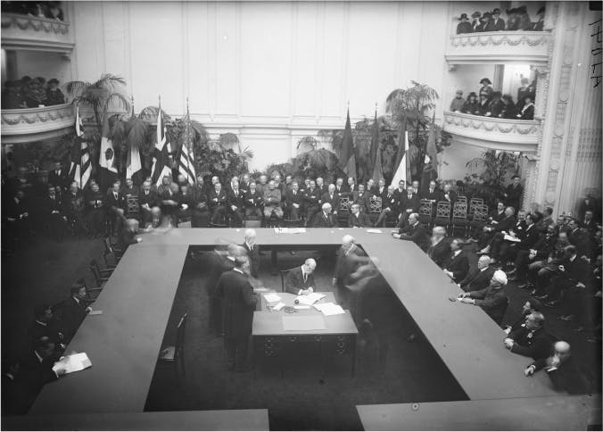 Men sit around a U-shaped table. A group of men sit at a table in the middle and sign some papers. Other men sit around the room and in balconies. Multiple flags are posted at the back of the room.