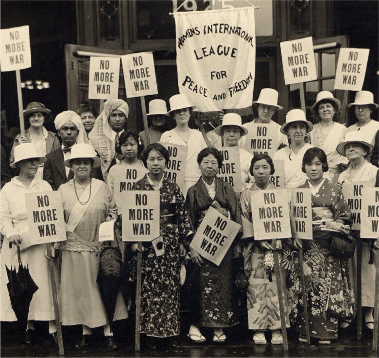 A group of women of different ethnicities and cultural dress stand together and hold signs that read No More War.