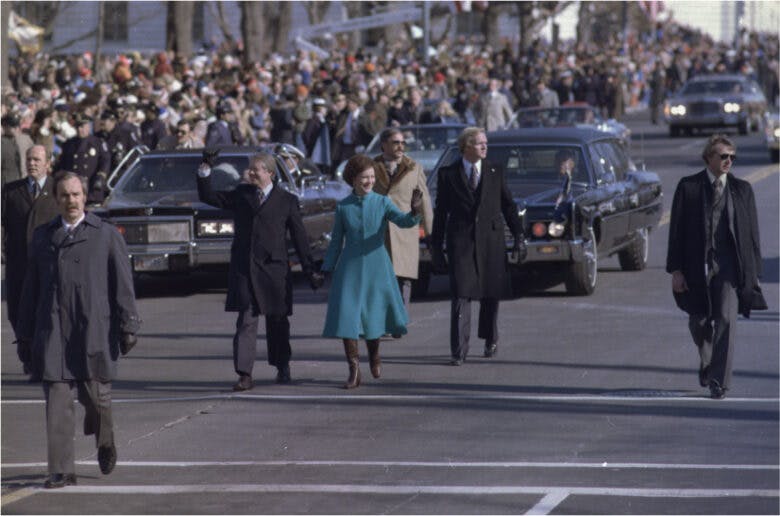 Jimmy Carter and his wife walk on the street in a parade. Cars follow them. A crowd lines the side of the street.