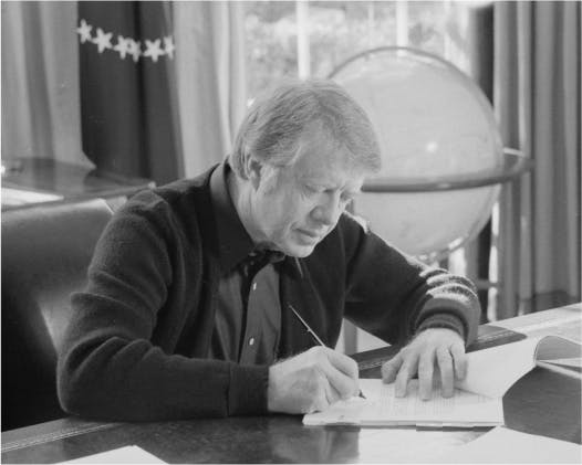 Jimmy Carter sits at a desk and signs a document.
