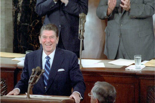 Ronald Reagan stands behind a podium. George H. W. Bush and Tip O'Neill stand behind him and clap. An American flag hangs behind them.